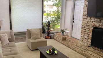 Un salon comprenant un canapé, une table basse et des poufs bleus. On y trouve une cheminée avec un manteau en pierre et un téléviseur fixé au mur.