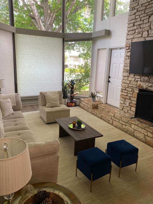 A living room with a sofa, coffee table, and blue ottomans. There is a fireplace with a stone surround and a TV mounted on the wall.