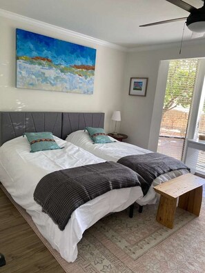 A bedroom with two beds, a ceiling fan, a painting, and a window with blinds.