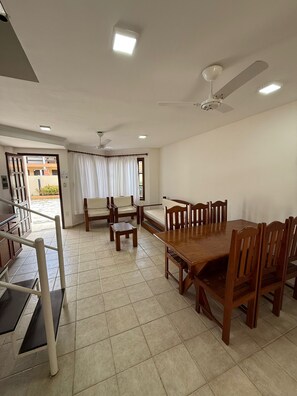 A dining area with a wooden table and chairs, a ceiling fan, and a view through a window with curtains. - Village dos bosques 43 (Porto Seguro)