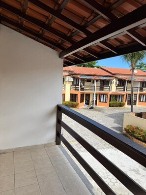 A covered balcony with a tiled floor, a wooden railing, and a view of a building with multiple balconies and a tiled roof. - Village dos bosques 43 (Porto Seguro)