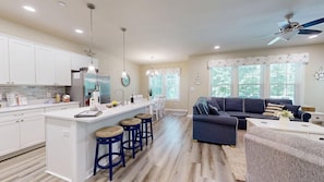 A modern kitchen with white cabinets, a central island, blue bar stools, and a dark blue sofa in a living area with large windows. - The Foxes Den (Ocean Pines)