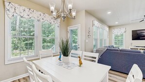 Un coin repas avec une table blanche, des chaises et un lustre. On aperçoit un canapé et un téléviseur à l’arrière-plan.