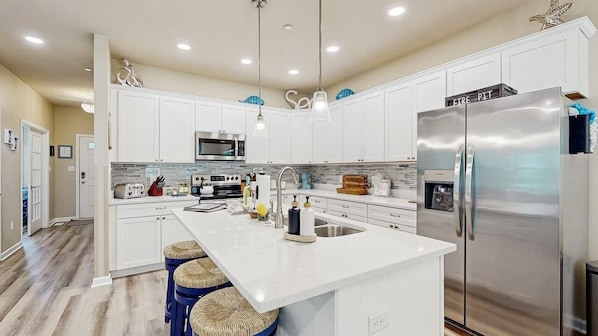 A modern kitchen with white cabinets, stainless steel appliances, and a central island with stools. - The Foxes Den (Ocean Pines)