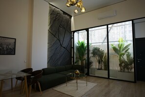 A modern living room with a glass dining table, a green sofa, and a large window with plants.