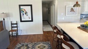 A modern kitchen with a central island, wooden chairs, a patterned rug, and framed artwork on the walls.