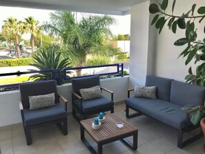 A balcony with two armchairs, a sofa, and a small table, overlooking a landscaped area with palm trees. - Cozy Apartment in Vilamoura Marina with Pool & Balcony (vilamoura)