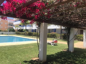 A pool area with a pergola covered in pink flowers, a lounge chair, and a building in the background. - Cozy Apartment in Vilamoura Marina with Pool & Balcony (vilamoura)