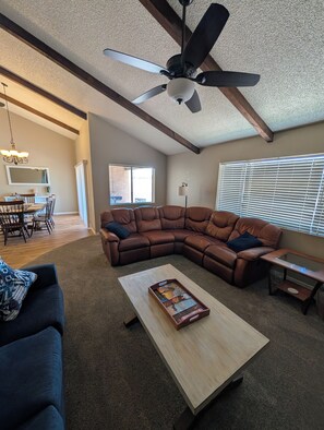 A living room with a sectional sofa, a coffee table, a ceiling fan, and a dining area with a chandelier. - Beautiful 2-bedroom townhome on golf course in Mesa (mesa)