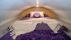 Um quarto de sótão pequeno, com teto inclinado, colcha roxa e manta de tricô.