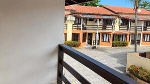 En overdækket balkon med flisegulv, trægelænder og udsigt til en bygning med flere balkoner og et fladt tag.