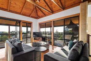 Een ruime woonkamer met een houten plafond, grote schuifdeuren, een zithoek, een salontafel en een televisie.