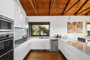 Een moderne keuken met witte kasten, roestvrijstalen apparatuur, een houten plafond en een raam met uitzicht op groen.