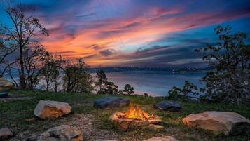 Una fogata rodeada de rocas y árboles, con vistas a un cuerpo de agua y un horizonte urbano a lo lejos.