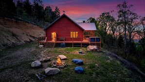Une cabane rouge dotée d’une terrasse, d’un foyer et offrant une vue sur la forêt environnante.
