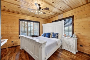 A bedroom with a large bed, a ceiling fan, wooden walls, and a window with blinds.