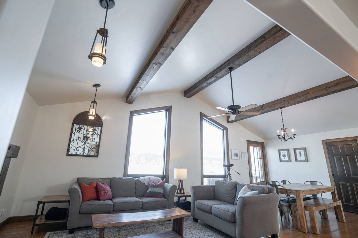 Un vaste salon doté d’un haut plafond, de poutres apparentes en bois, d’un canapé gris, d’une table basse en bois et d’un coin repas avec une table et des chaises en bois.
