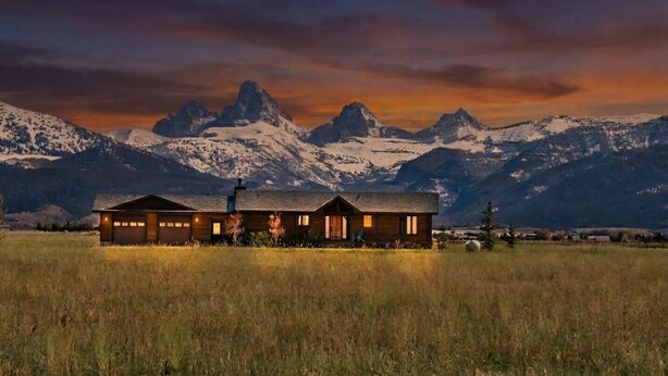 Una cabaña con garaje, rodeada por un campo, con montañas al fondo.