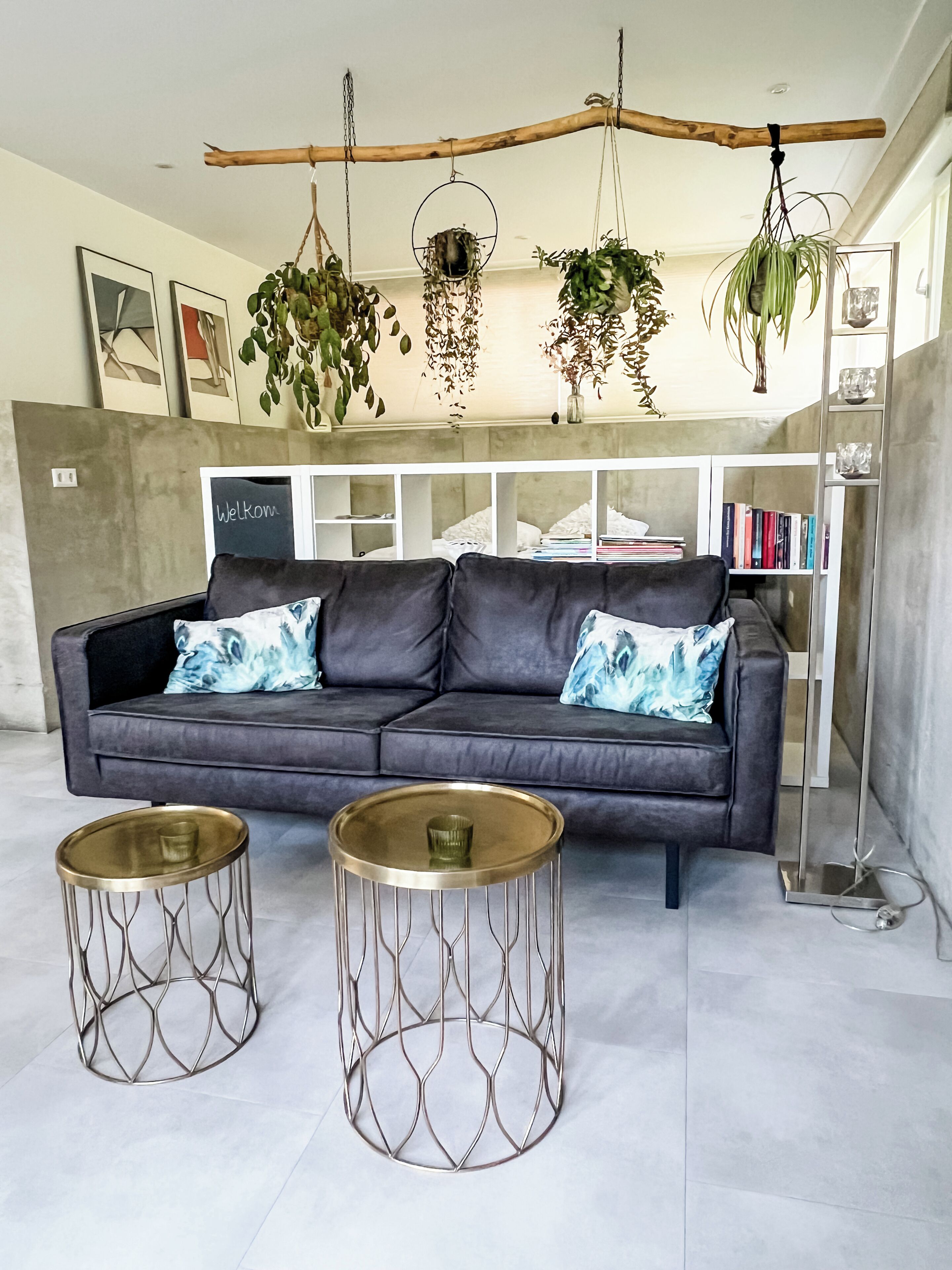 Ein modernes Wohnzimmer mit einem grauen Sofa, zwei metallischen Beistelltischen und hängenden Pflanzen.