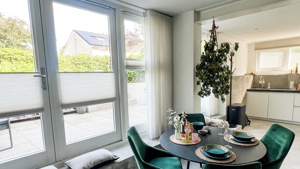 A dining area with a table set for four, green chairs, a hanging plant, and a view through large windows. - Studio Margriet - coming home to Bergen (Bergen)