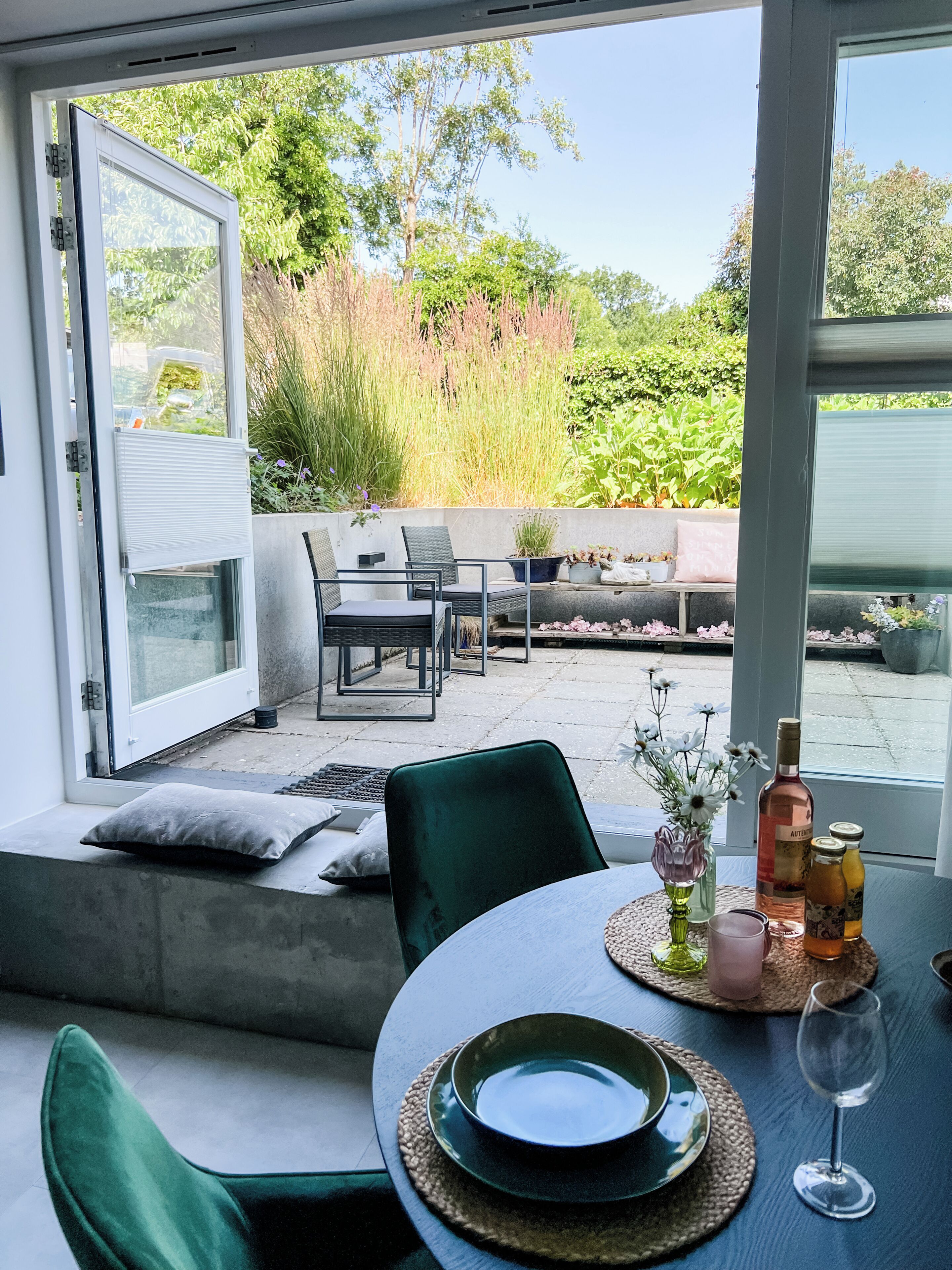 Een open glazen schuifdeur leidt naar een terras met een tafel voor twee personen, een bankje en uitzicht op groen.