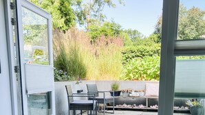 An open sliding glass door leading to an outdoor seating area with a table set for two, a bench, and a view of greenery. - Studio Margriet - coming home to Bergen (Bergen)