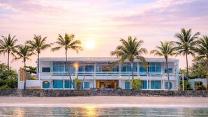 Una propiedad frente a la playa con un edificio de dos pisos, palmeras y vista al mar.