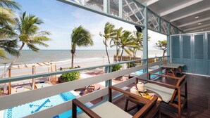 Un balcón con vistas a la playa y al océano, sillas de madera, una mesa pequeña con un pastel y una piscina.