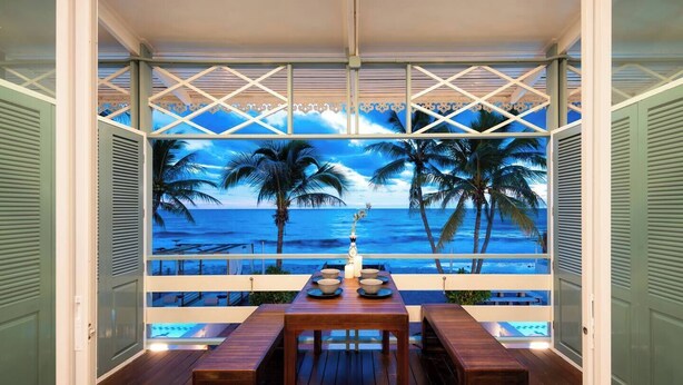 Una terraza de madera con una mesa y bancos, con vistas a una playa con palmeras y el océano.