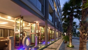 A modern hotel exterior with glass doors, a water feature, and palm trees. - 2BR Comfort Pool Access Bangtao & Nightlife C178 (Chang Wat)