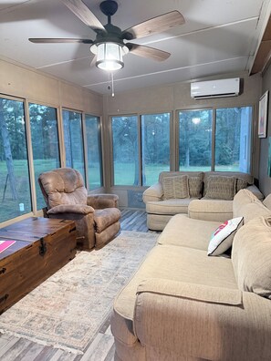 Ein geräumiges Wohnzimmer mit großen Fenstern, einem Deckenventilator und einem Couchtisch aus Holz.
