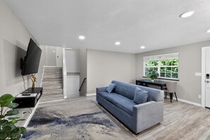Una sala de estar moderna con un sofá gris, un televisor de pantalla plana, una escalera y un ventanal con vistas a zonas verdes.