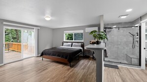 A modern bedroom with a bed, a bedside table, a potted plant, a glass shower enclosure, and a view of the outdoors.