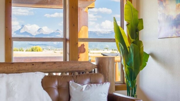 Ein Zimmer mit Blick auf Berge, durch große Fenster sichtbar, mit einem braunen Ledersessel, einem weißen Kopfkissen und einer großen Grünpflanze.