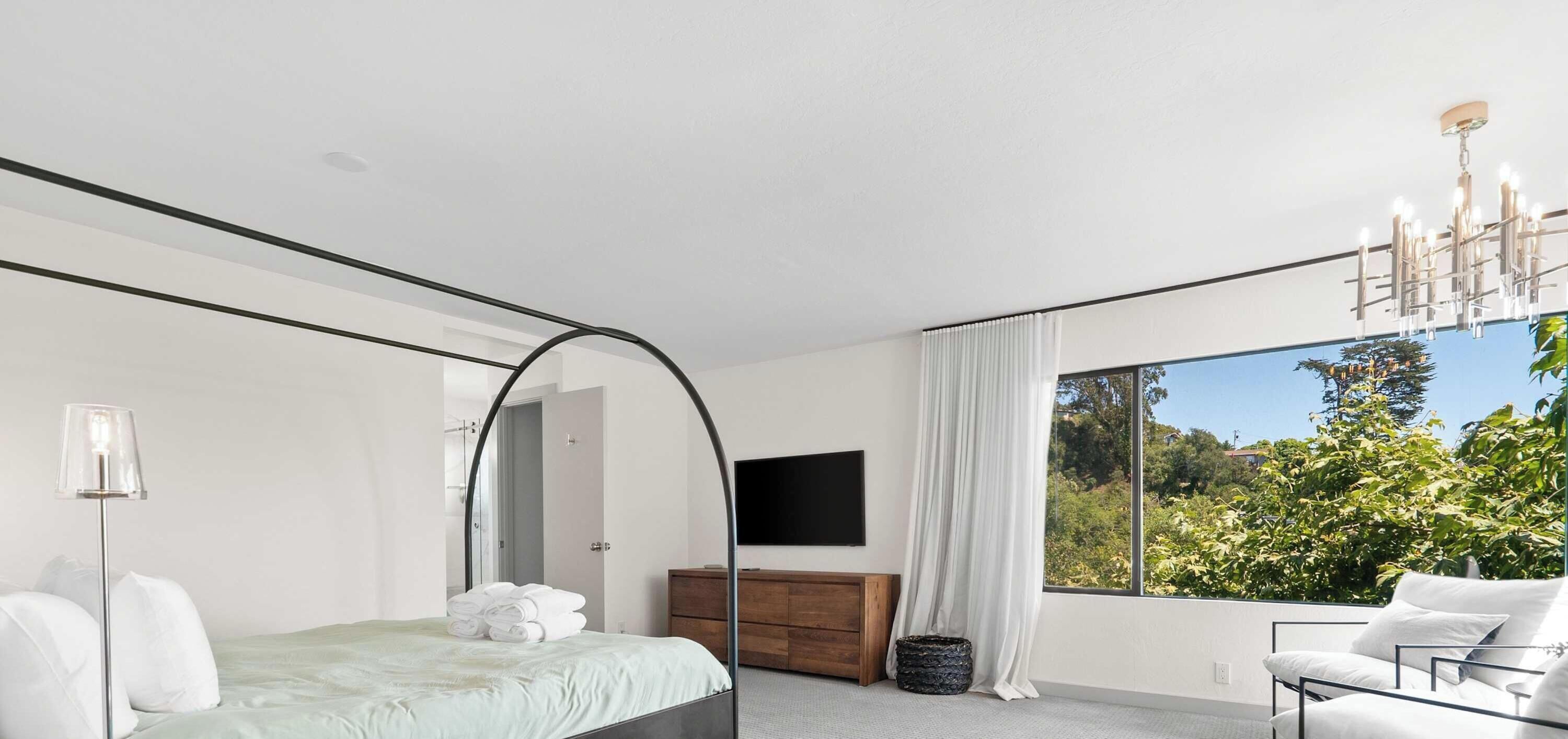 Ein modernes Schlafzimmer mit Bett, Kommode, Fernseher und einem großen Fenster mit Blick auf Grünflächen.