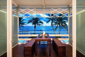 Eine Holzterrasse mit Tisch und Bänken, die auf einen Strand mit Palmen und den Ozean blickt.
