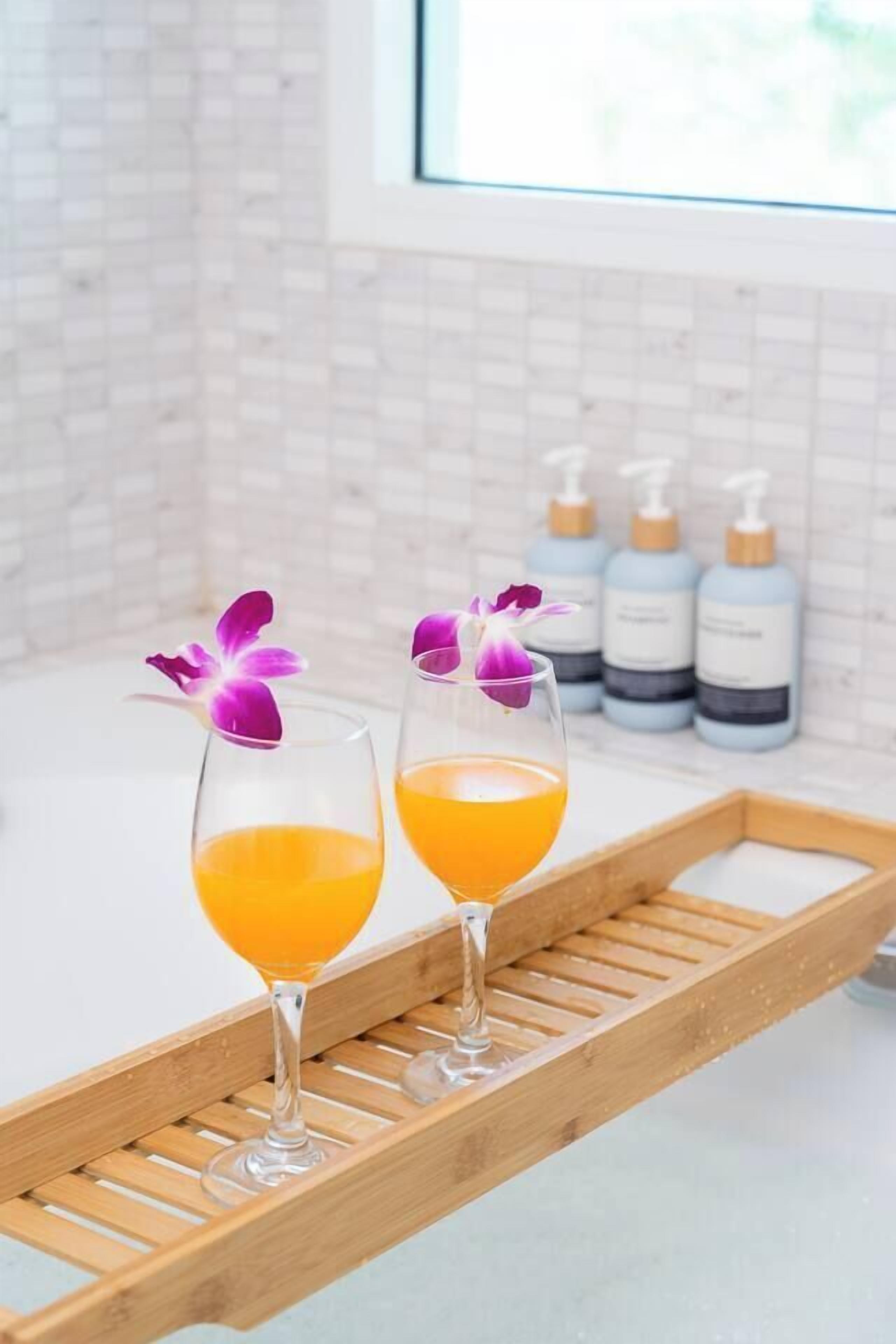 Dos vasos de jugo de naranja sobre una bandeja de madera con flores moradas, en una habitación con paredes de azulejos blancos y botellas al fondo.
