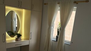 Une chambre à coucher avec un lit, une coiffeuse avec un miroir, une plante en pot et une fenêtre avec des rideaux.