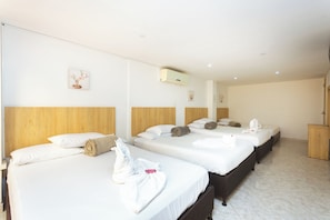 Habitación de hotel con cuatro camas, cada una con ropa de cama y toallas blancas, cabeceros de madera y suelo de baldosas blancas.