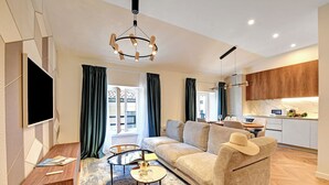 Una sala de estar moderna con un sofá, una mesa de centro y un televisor. Hay una zona de cocina con armarios y una mesa de comedor.
