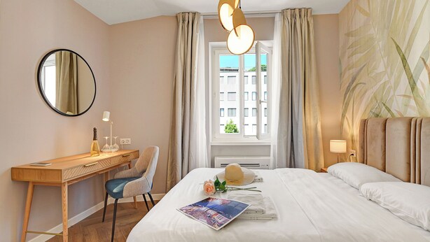 Habitación de hotel con un escritorio de madera, una silla, una cama con ropa blanca, un espejo redondo y una ventana con cortinas.