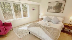 Una camera da letto con un letto, comodini, un tavolino, una finestra, una sedia rosa e un tappeto.