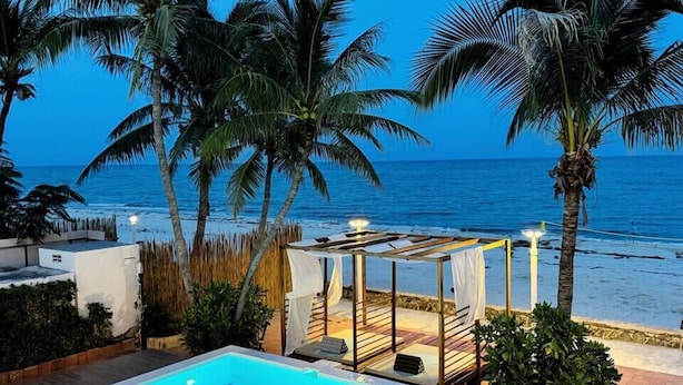 Ein Swimmingpool mit blauem Fliesendesign, umgeben von Liegestühlen und einer überdachten Sitzgruppe, mit Palmen und Blick auf den Ozean.