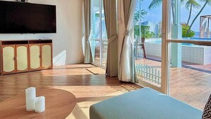 Ein Zimmer mit Flachbildfernseher, einem Holzschrank, einem runden Tisch mit Kerzen, einer Couch und großen, mit Vorhängen versehenen Fenstern.