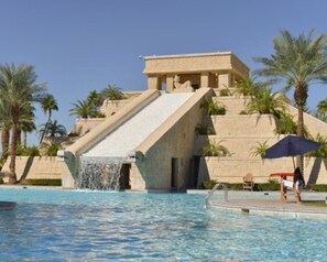 Une piscine agrĂ©mentĂ©e dâune cascade, de palmiers et dâun bĂątiment Ă Ă©tages.