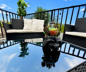 Ein Glastisch mit einer schwarzen Vase, eine Rattan-Couch mit Kissen und ein Topf mit Sukkulenten.
