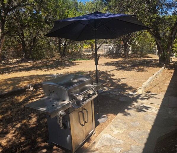 An outdoor grill setup with a canopy umbrella, surrounded by trees and a stone pathway.