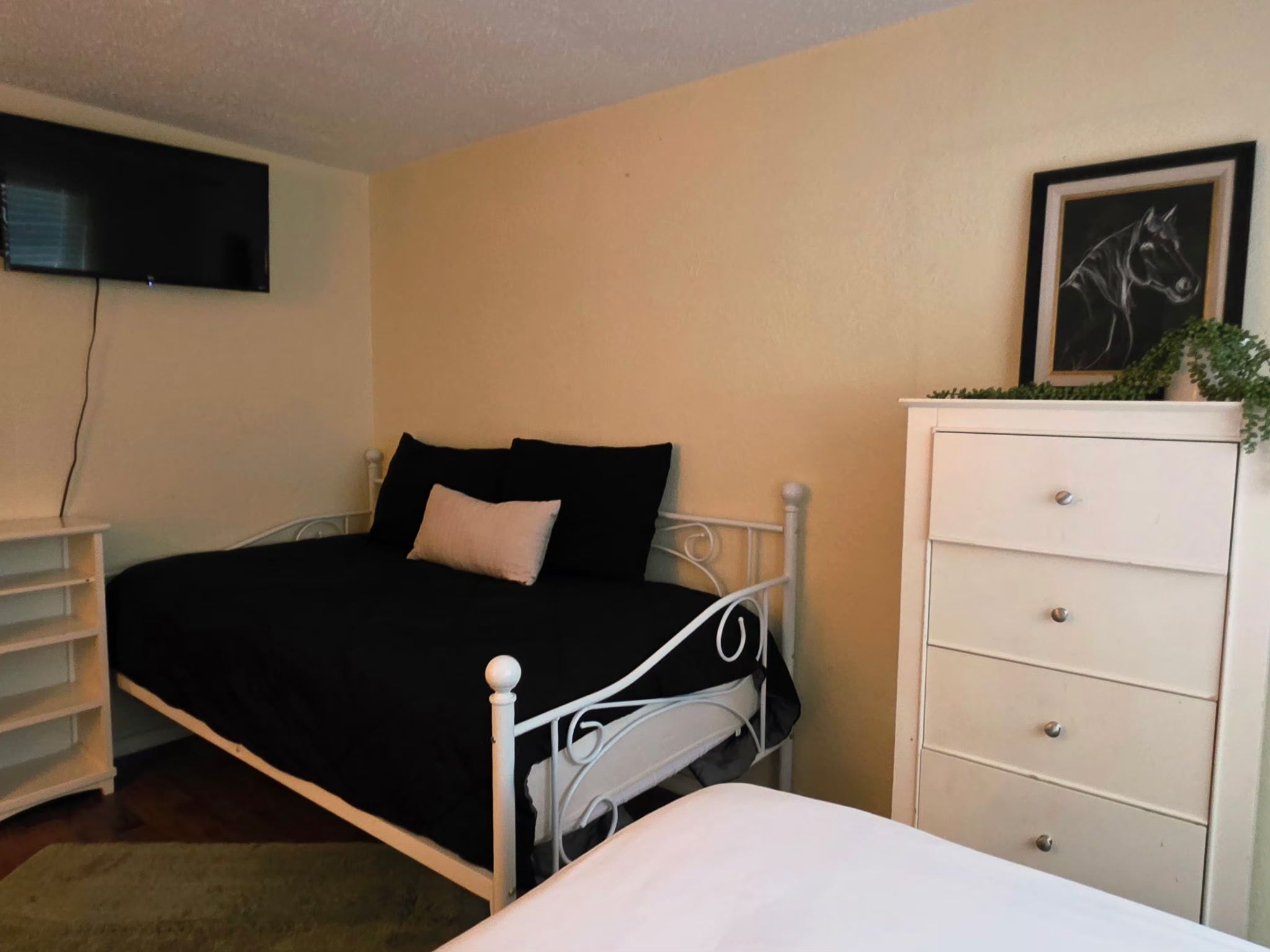 Ein Schlafzimmer mit einem Bett, einer Kommode, einem an der Wand befestigten Fernseher und einer Pflanze auf der Kommode.