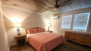 Une chambre avec un plancher en bois, un lit recouvert d’une couvre-lit à motifs, deux tables de chevet avec des lampes, une commode et un ventilateur de plafond.