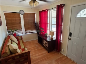 A room with a wooden floor, a sofa, a table with a lamp, a potted plant, and a door with a window.